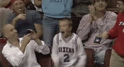 A Man In A Sixers Jersey Is Sitting In A Crowd Of People Watching A Basketball Game . GIF