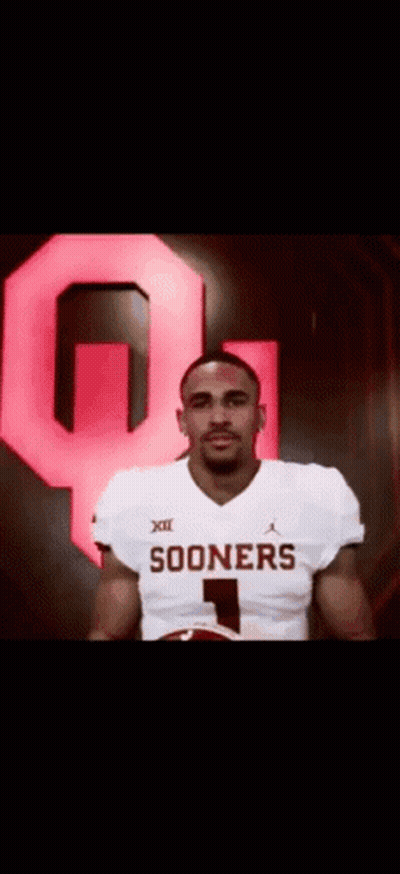 A Man In A Sooners Jersey Is Holding A Football In Front Of A Large Red Q . GIF