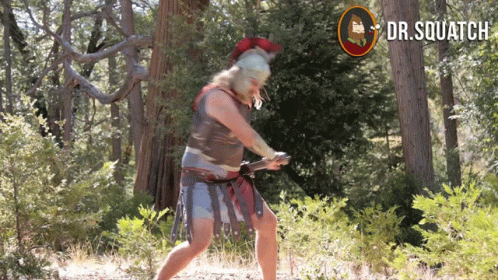 A Man In A Spartan Costume Is Holding A Sword In The Woods With A Dr.squatch Logo Behind Him GIF