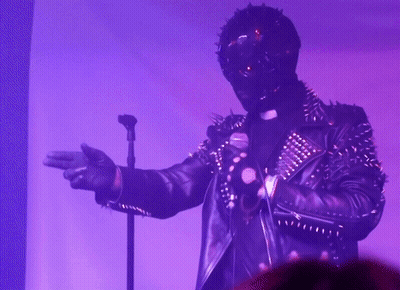 A Man In A Spiked Leather Jacket Is Singing Into A Microphone In A Dark Room GIF