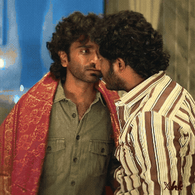 A Man In A Striped Shirt Is Kissing Another Man In A Green Shirt GIF