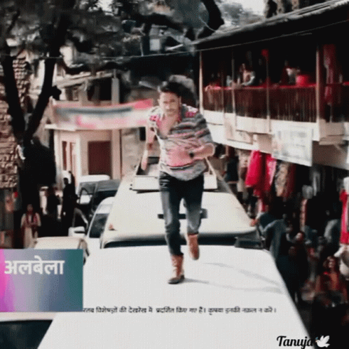A Man In A Striped Shirt Is Running Down A Street In Front Of A Sign That Says Tanuja GIF