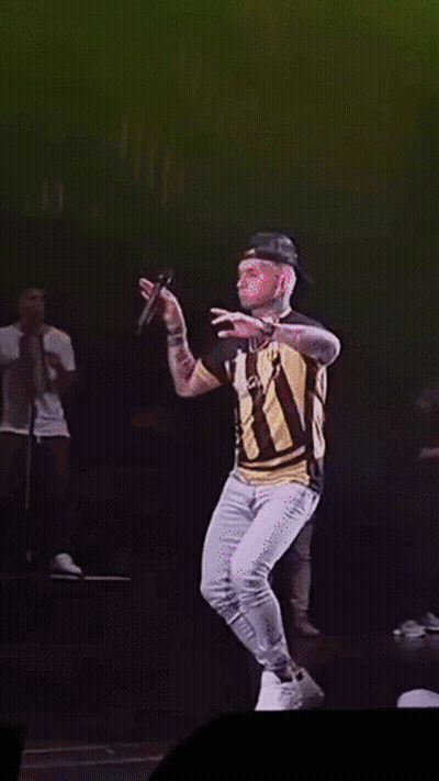 A Man In A Striped Shirt Is Singing Into A Microphone On Stage GIF