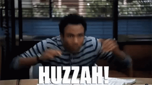 A Man In A Striped Shirt Is Sitting At A Desk With The Words Huzzah Written On The Table . GIF