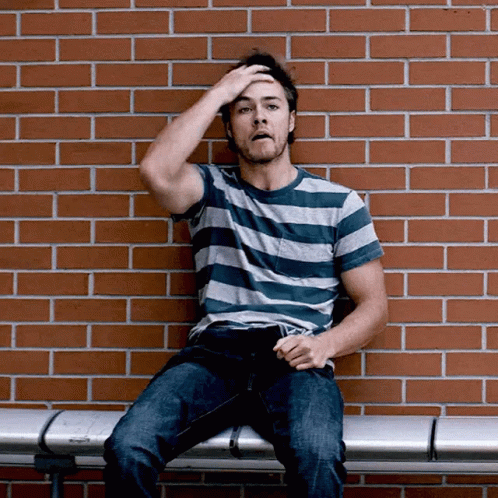 A Man In A Striped Shirt Is Sitting On A Bench In Front Of A Brick Wall GIF