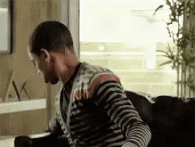 A Man In A Striped Shirt Is Sitting On A Couch Looking Out A Window . GIF