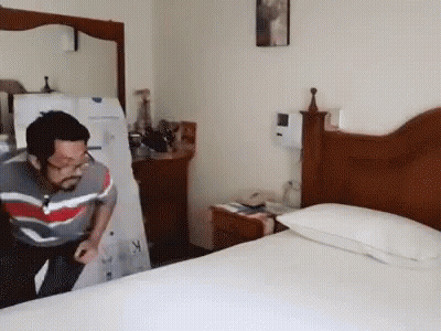 A Man In A Striped Shirt Is Standing In A Bedroom Next To A Bed GIF