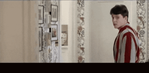 A Man In A Striped Shirt Is Standing In A Hallway Looking At A Wall With Pictures On It . GIF