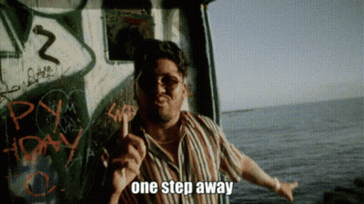 A Man In A Striped Shirt Is Standing In Front Of A Wall With Graffiti And Says One Step Away GIF