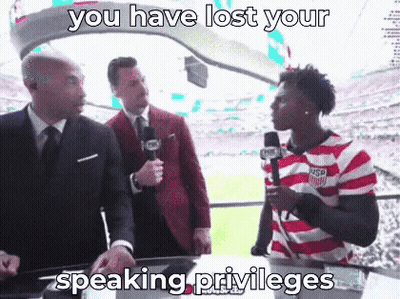 A Man In A Striped Shirt Is Talking Into A Microphone While Two Men In Suits Look On . GIF