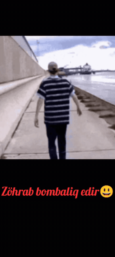 A Man In A Striped Shirt Is Walking Down A Sidewalk Next To The Water . GIF
