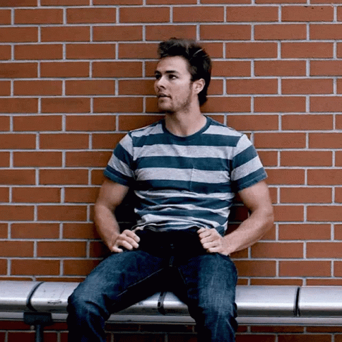 A Man In A Striped Shirt Sits On A Bench In Front Of A Red Brick Wall GIF