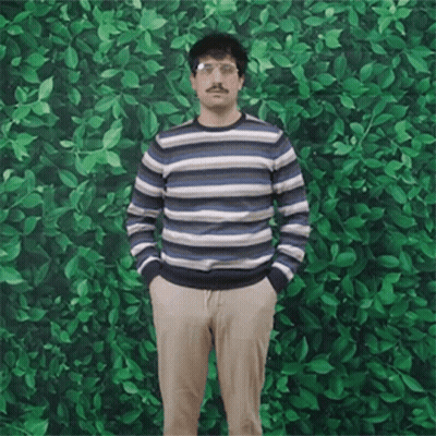 A Man In A Striped Sweater Stands With His Hands In His Pockets In Front Of A Green Wall GIF