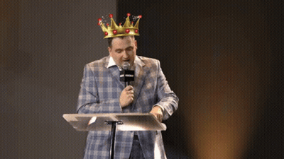 A Man In A Suit And A Crown Is Standing At A Podium With A Microphone . GIF