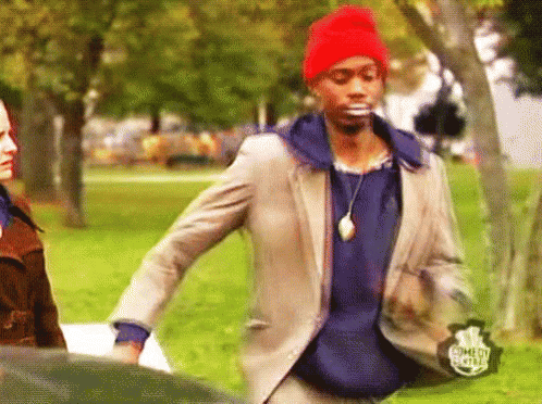 A Man In A Suit And A Red Hat Is Standing In A Park With A Comedy Central Logo On His Jacket GIF