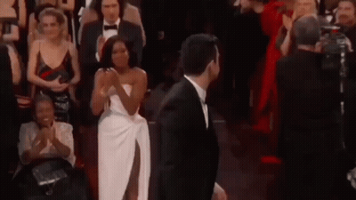 A Man In A Suit And A Woman In A White Dress Are Standing In Front Of A Crowd Of People . GIF