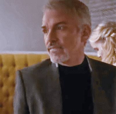 A Man In A Suit And Black Turtleneck Is Standing In Front Of A Woman . GIF