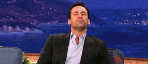 A Man In A Suit And Blue Shirt Is Sitting In A Chair With His Eyes Closed And His Mouth Open . GIF