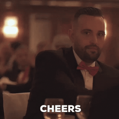 A Man In A Suit And Bow Tie Is Holding A Glass Of Wine And The Word Cheers Is Behind Him GIF