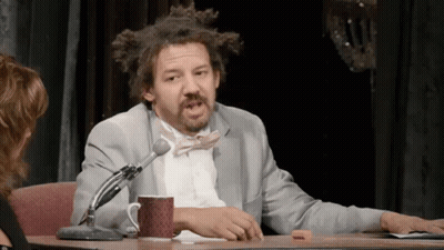 A Man In A Suit And Bow Tie Is Sitting At A Table With A Microphone And A Cup Of Coffee . GIF