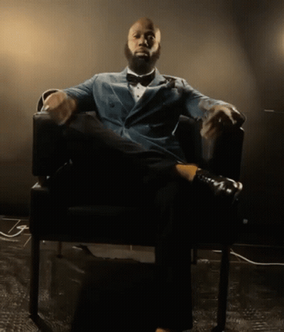 A Man In A Suit And Bow Tie Is Sitting In A Chair With His Legs Crossed GIF