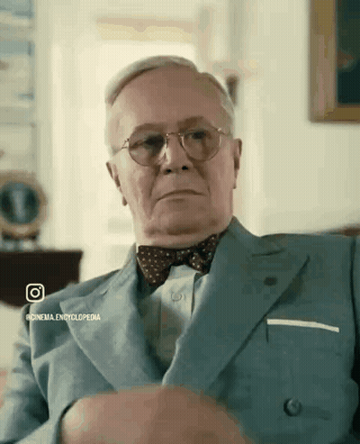 A Man In A Suit And Bow Tie Is Sitting On A Couch GIF