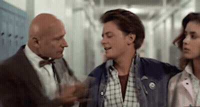 A Man In A Suit And Bow Tie Is Talking To A Young Man In A Plaid Shirt In A Hallway . GIF
