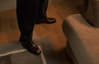 A Man In A Suit And Brown Shoes Is Walking Down Stairs In A Living Room . GIF