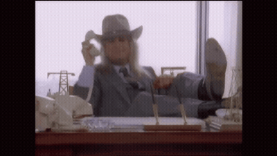 A Man In A Suit And Cowboy Hat Is Sitting At A Desk With His Feet Up Talking On A Cell Phone . GIF