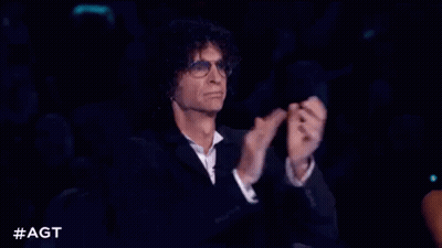 A Man In A Suit And Glasses Is Sitting In A Dark Room Clapping His Hands . GIF