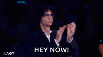 A Man In A Suit And Glasses Is Sitting In A Dark Room With His Hands In The Air . GIF