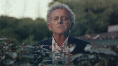A Man In A Suit And Glasses Is Standing In Front Of A Bush With His Eyes Closed GIF