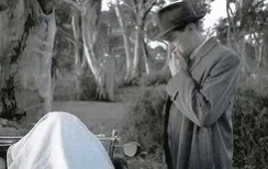 A Man In A Suit And Hat Is Smoking A Cigarette While Standing Next To A Body . GIF
