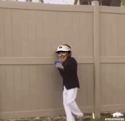 A Man In A Suit And Hat Is Standing In Front Of A Fence Holding A Cup . GIF