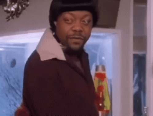 A Man In A Suit And Hat Is Standing In Front Of A Lava Lamp And Looking At The Camera . GIF