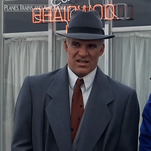 A Man In A Suit And Hat Is Standing In Front Of A Neon Sign That Says Planes Trains And Automobiles GIF