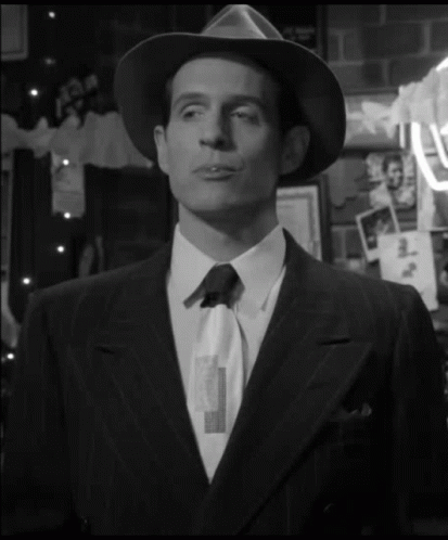 A Man In A Suit And Hat Is Standing In Front Of A Wall With Pictures GIF