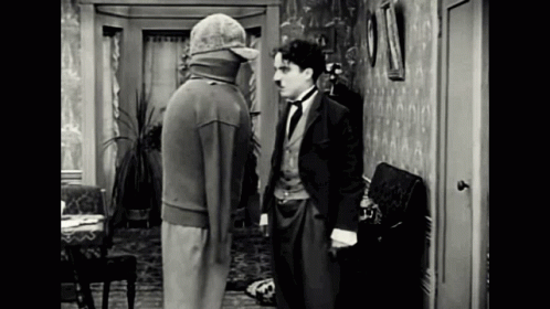 A Man In A Suit And Hat Is Standing Next To Another Man In A Room . GIF