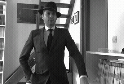 A Man In A Suit And Hat Stands In Front Of A Bookshelf With Magazines On It GIF