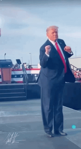 A Man In A Suit And Red Tie Stands On A Stage With His Fist In The Air GIF