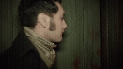 A Man In A Suit And Scarf Is Standing In Front Of A Wooden Wall And Looking At The Camera . GIF