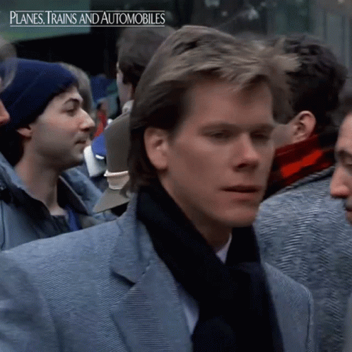 A Man In A Suit And Scarf Stands In A Crowd With Planes Trains And Automobiles Written Above Him GIF