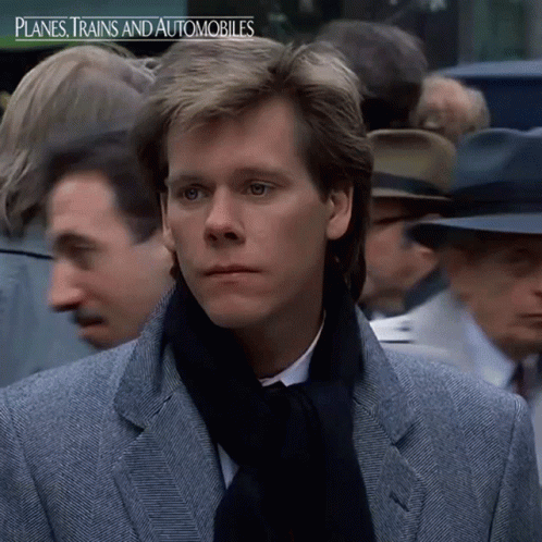 A Man In A Suit And Scarf Stands In Front Of A Crowd Of People With The Words Planes Trains And Automobiles Below Him GIF