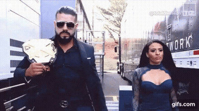 A Man In A Suit And Sunglasses Is Holding A Wrestling Championship Belt Next To A Woman In A Blue Dress . GIF
