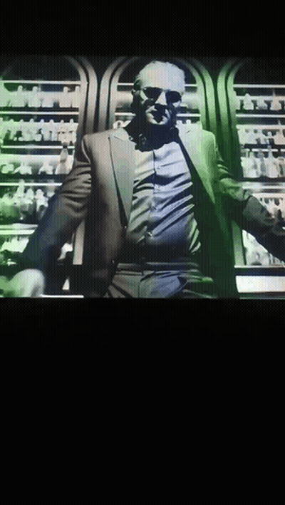 A Man In A Suit And Sunglasses Is Standing In Front Of A Display Of Bottles GIF