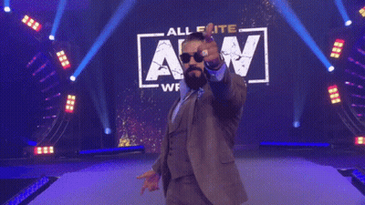 A Man In A Suit And Sunglasses Is Standing In Front Of A Sign That Says Wrestling Saturday Night Dynamite GIF