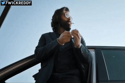 A Man In A Suit And Sunglasses Smoking A Cigarette In A Car With The Twitter Username 7Wickreddy Below Him GIF