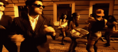A Man In A Suit And Sunglasses Stands In Front Of A Group Of People Dancing GIF