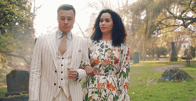 A Man In A Suit And Tie And A Woman In A Floral Dress Are Walking In A Park . GIF