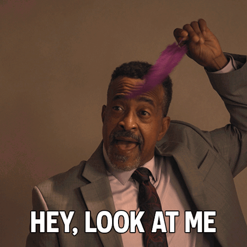 A Man In A Suit And Tie Holds A Feather In His Hair And Says " Hey Look At Me " GIF